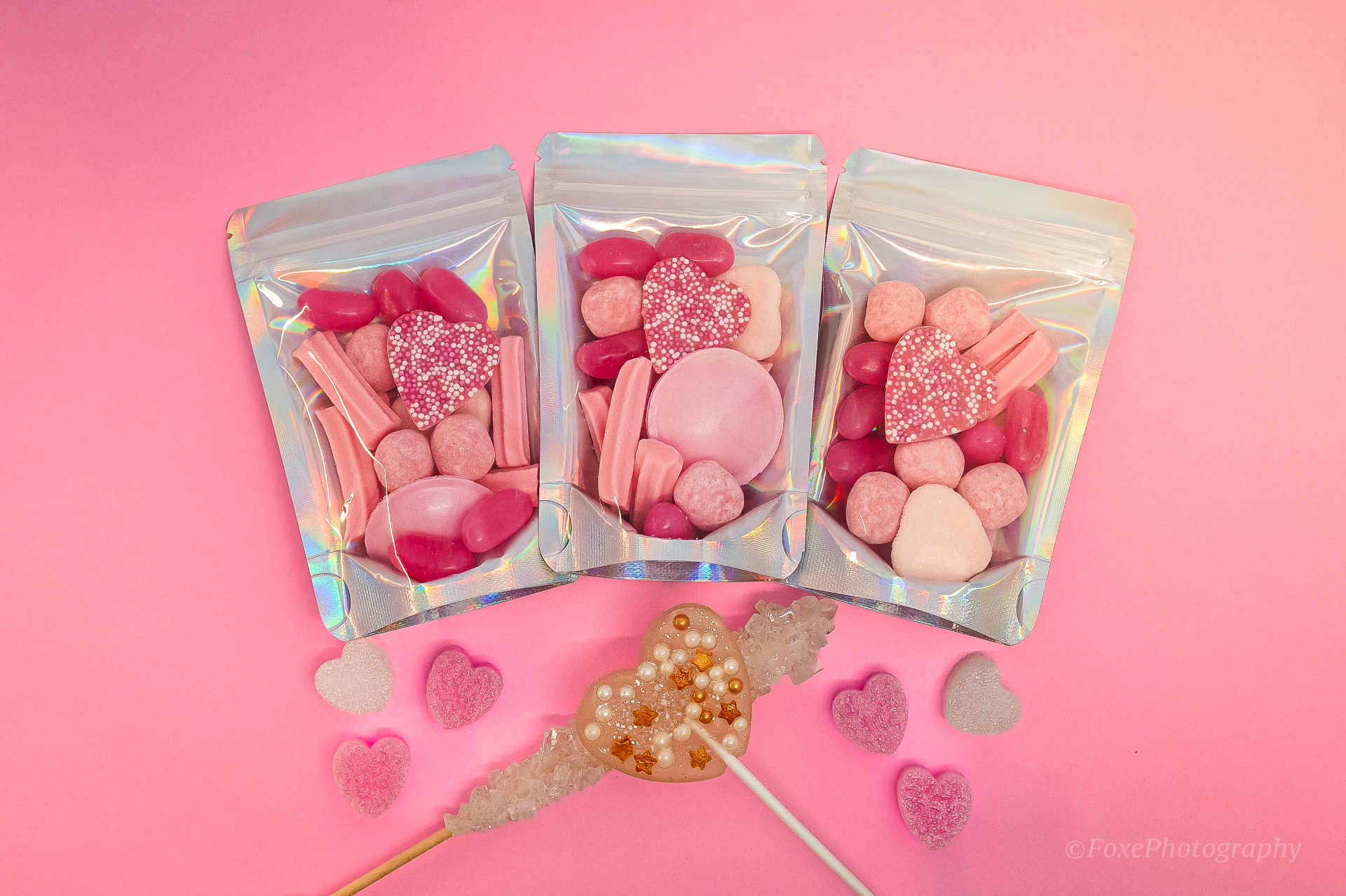 Three holographic favour bags filled with pink lollies, jelly beans, musk sticks and heart lollipops on a pink backdrop