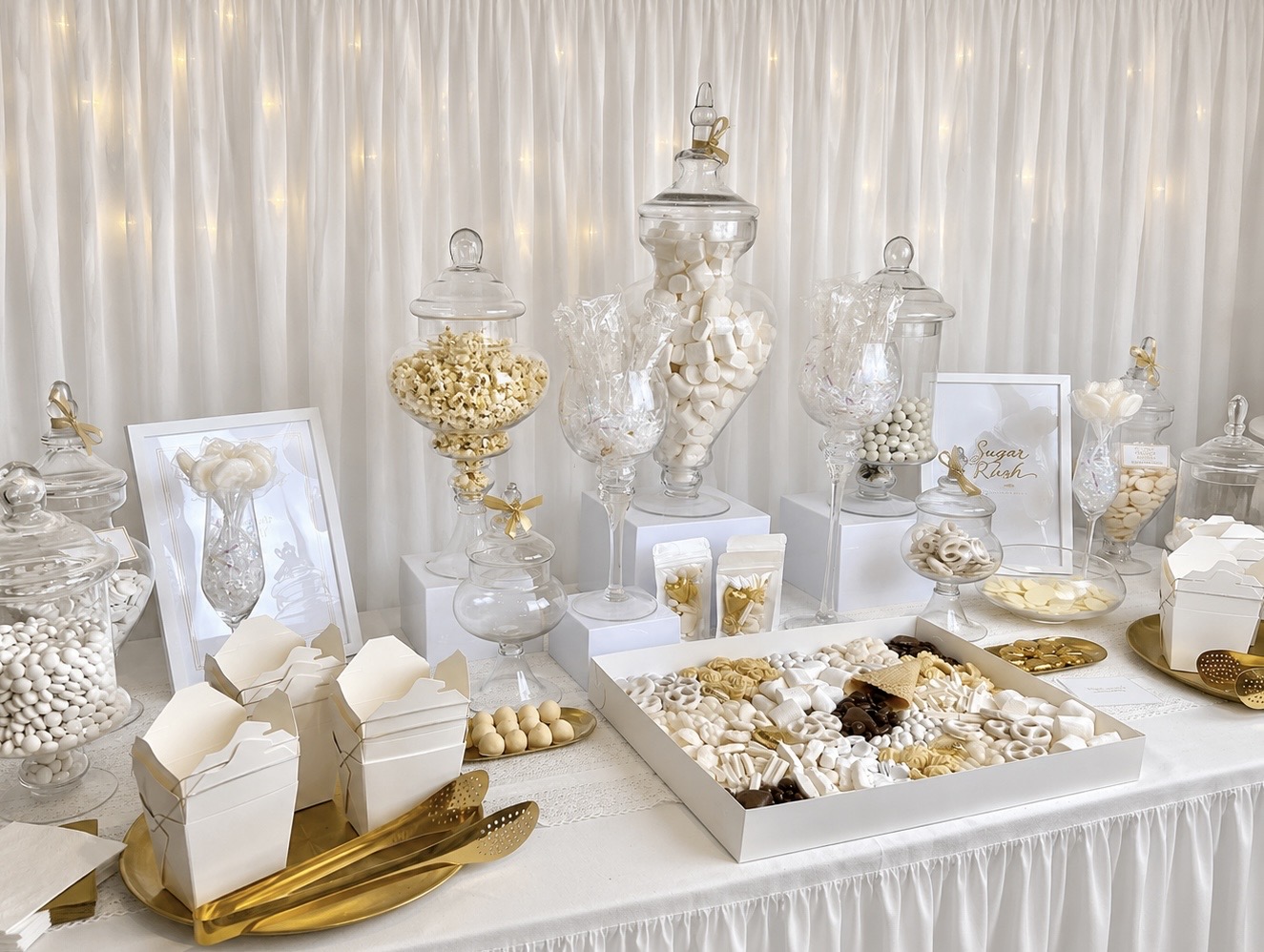 Elegant white and gold dessert bar with apothecary jars, popcorn, marshmallows and sweets on a draped white backdrop