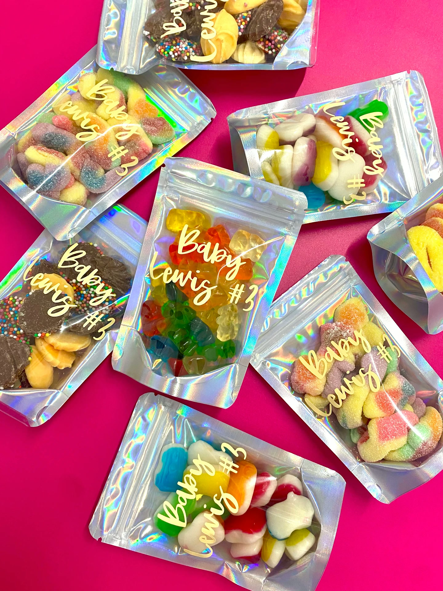 Holographic silver favour bags with gold 'Baby Lewis' script lettering, each filled with different colour-coordinated lollies on a hot pink background