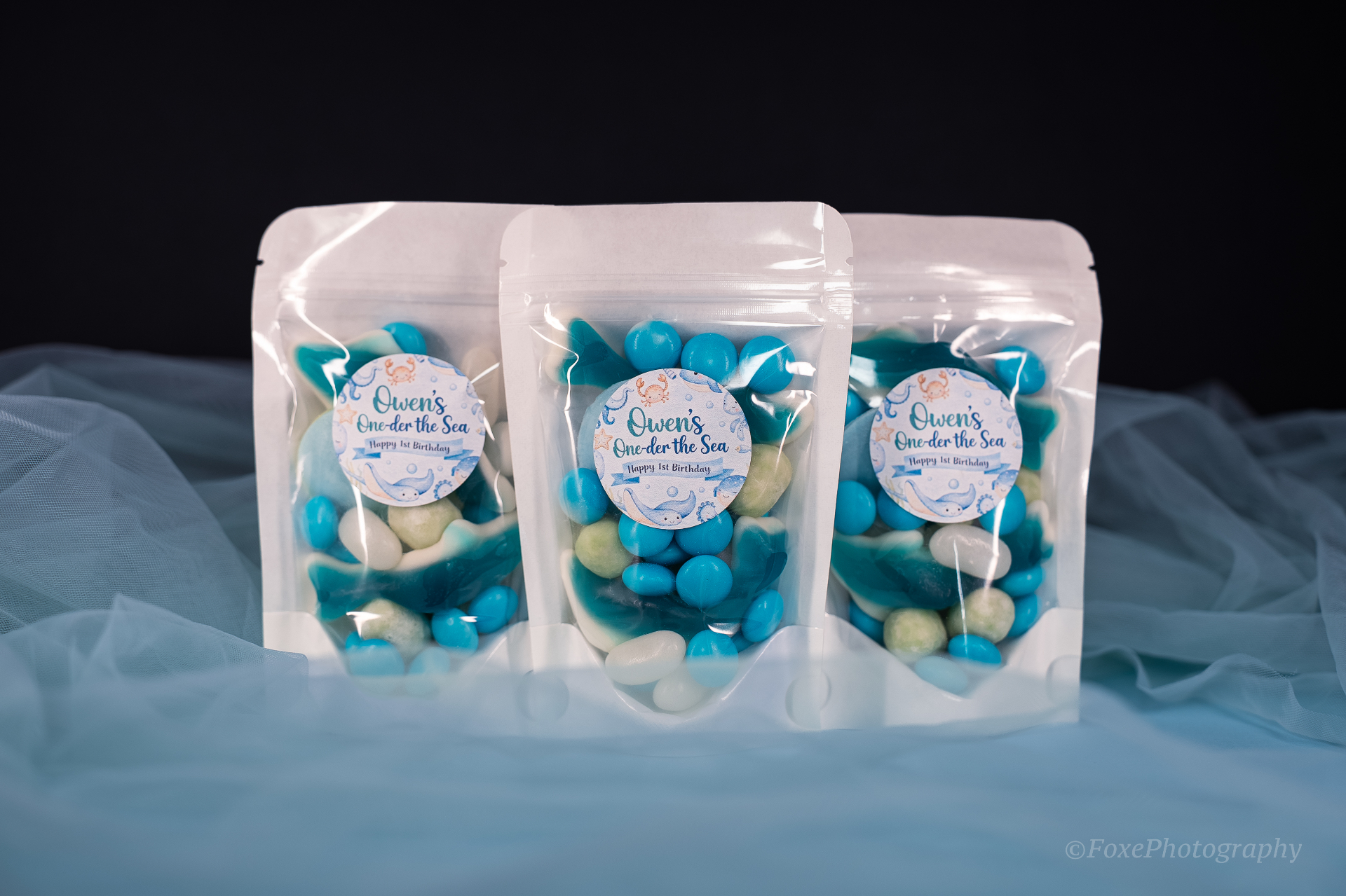Three custom Under the Sea themed lolly bags filled with blue, white and green sweets, tagged 'Owen's One-der the Sea Happy 1st Birthday' on soft blue tulle